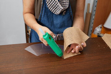 close up of woman hands gluing sackcloth to plastic with glue gun on workshop table