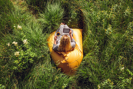 One woman person reading an ebook electronic book and sit on yellow beanbag in wild nature in summer. Above view.