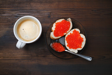 Sandwiches with red caviar, coffee cup, plate and spoon on wooden background. Flat lay.