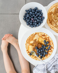 Pancakes, blueberries and honey on a white table on a gray concrete background. Maslenitsa concept and summer time. Kid's legs on the background, child eating pancakes, children's summer menu