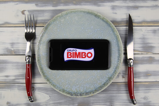 Viersen, Germany - May 9. 2021: Closeup of mobile phone on plate and wood table, cutlery with logo lettering of mexican grupo bimbo food company (focus on center)