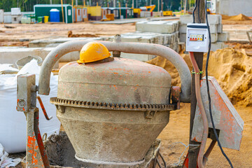 An industrial concrete mixer at a construction site with an orange worker's helmet lying on it. Concrete preparation.