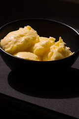 Rich creamy textured butter closeup view from the front. The golden yellow butter served in black ceramic bowl, placed on black background, lit up by harsh sunlight shines,glows and casts deep shadows