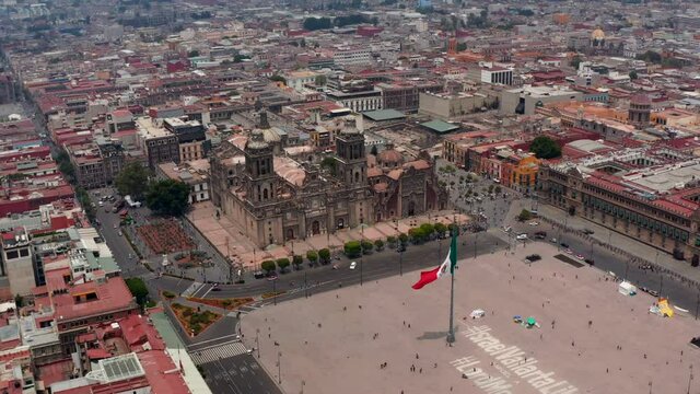Forwards fly over Plaza de la Constitucion with large state flag. Aerial view of Catedral Metropolitana de la Ciudad de Mexico. Mexico City, Mexico.