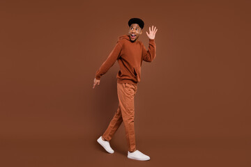 Fototapeta na wymiar Full length profile photo of funky short hairdo young guy wave empty space dress sportswear trousers sneakers isolated on brown color background