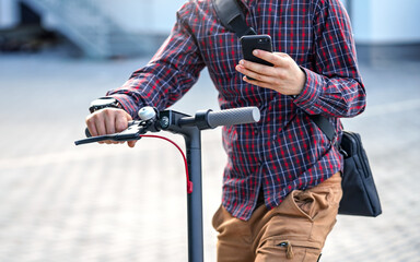 Young man wearing shirt leaning on electric scooter handlebar, holding mobile smartphone in his...
