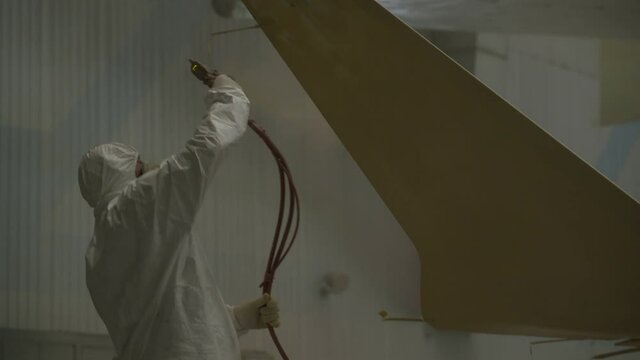 A worker in a white protective suit with a hood and a respirator paints the wing of an airplane with paint from a bullet gun.