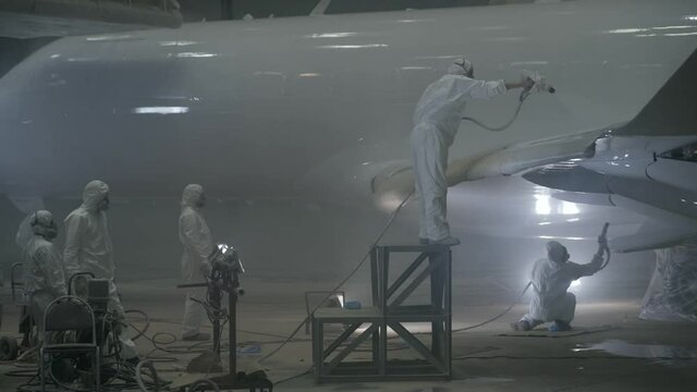 A group of workers in white protective suits and respirators stands next to the plane. The aircraft is being painted white.