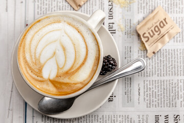Elegant modern close-up overhead shot of a cappuccino with a beautiful design in the foam.  Saucer below with a spoon and a blackberry.  Ripped brown sugar packet atop a daily newspaper. 