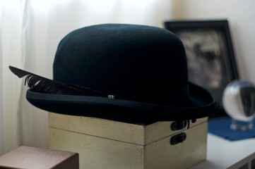 A man's black felt bowler hat with feather sits on top of wooden box in front of window.