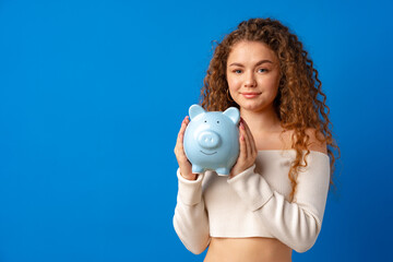 Fototapeta premium Curly-haired young woman holding piggy bank against blue background
