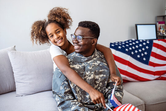 Happy little girl daughter with American flag hugging father in military uniform came back from US army, view of dad male soldier reunited reunited with family at home