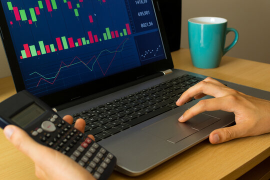 Young person holding a calculator, reviewing and investing in the stock market with technical indicator tools on his computer, with a cup to the side. Trading concept.