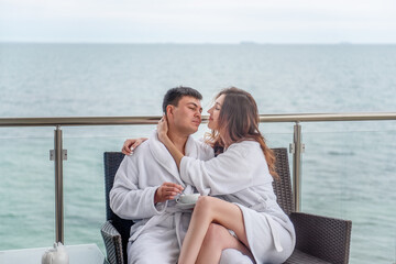 A couple in white bathrobes are sitting on an open terrace by the sea, drinking tea with lemon from cups. Lovers travel on vacation. Young man and woman hug each other. Newlyweds on their honeymoon