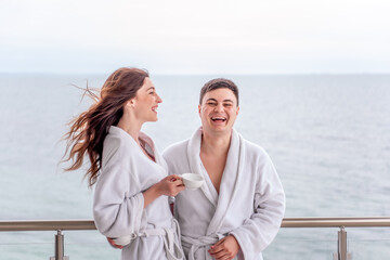Young loving couple in white terry robes drink tea from cups, the background of blue sea. Newlyweds...