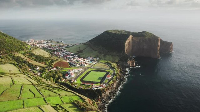 Aerial view of green island of Sao Jorge with rocky coastline, Azores, Portugal, Europe. Drone shot of village houses surrounded rural area. Football stadium located on cliffs at Atlantic ocean, 4k