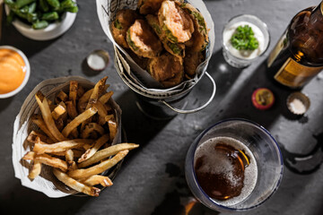 Overhead shot of bar food on a black slate surface.  One bucket holds fried pickles, one has French fries.  There is a beer bottle, a glass of beers well as a side of green beans, ranch dip and sauce.