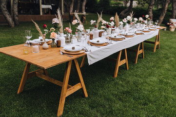 A beautiful wooden, long, decorated with flowers, reeds, empty dishes, plates, forks, knives, glasses with a white tablecloth stands on the green grass in the forest. Wedding banquet.
