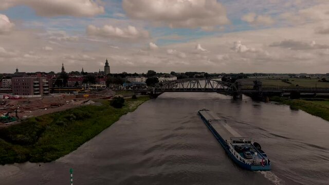 Aerial following large cargo vessel ship leaving wrinkles in the water of the river IJssel passing underneath steel draw bridge with train and historic Hanseatic city of Zutphen, The Netherlands.