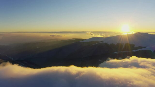 Cloud waves gently and slowly flow over the tops of snow-capped mountains covered with spruce forests at a cold sunset