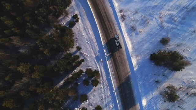 Modern white boom lift truck drives along empty snowy rural road past pine forest in winter evening bird eye view