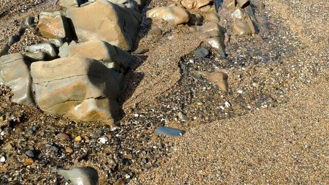 Close up of a small water stream along the beach that flows to the sea between some rocks. It has created a gap on the sand and lots of pebbles are seen below the water.