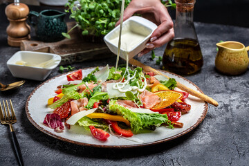 Pouring white sauce over plate of cooked salad with pieces of smoked salmon, lettuce, sun dried...