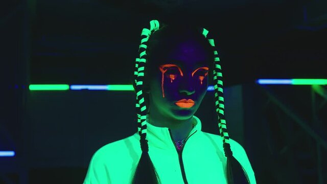 Portrait of female stylish artist inside dark place with neon lights .  Beautiful young girl with fluorescent colorful make-up and cloth in UV light . Portrait of woman under UV black light 