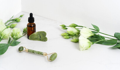Jade roller for face massage. Green gua sha on the marble white background with flat lay. Anti age, lifting and toning treatment, acupressure. Eustoma flowers. Serum brown bottle with pipette