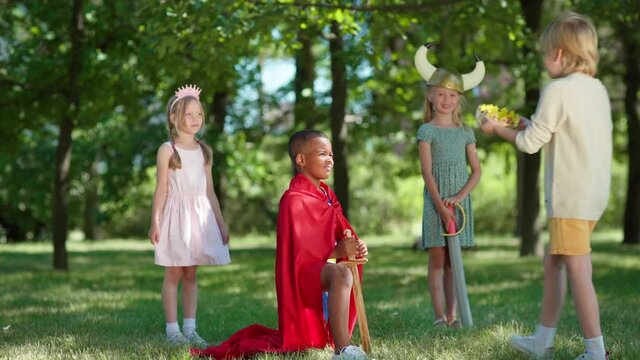 Diverse kids in costumes role playing in park. Black little boy in red cape being coronated kneeing on grass with sword in his hand. Friends applauding to him