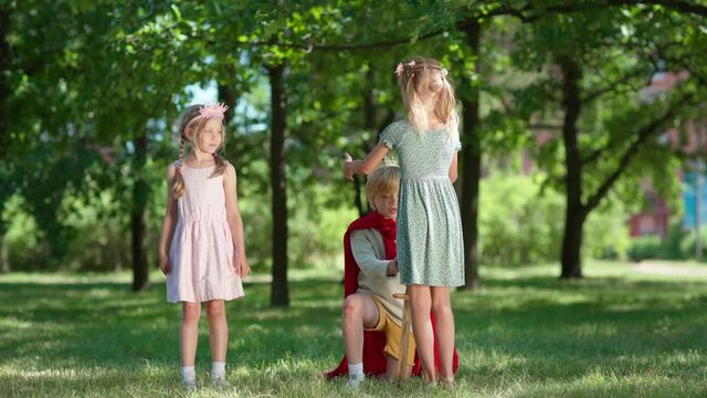 Kids in costumes role playing in park. Little boy in red cape being coronated kneeing on grass with sword in his hand. Girl putting crown on his head. Children applauding to king