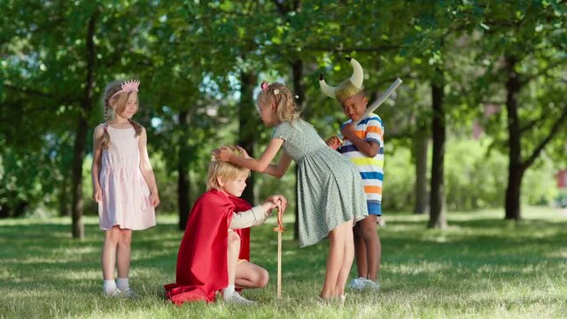 Diverse kids in costumes role playing in park. Boy being coronated kneeing on grass. Girl putting crown on is head. Happy child standing up and raising arm arm with sword. Children applauding to king