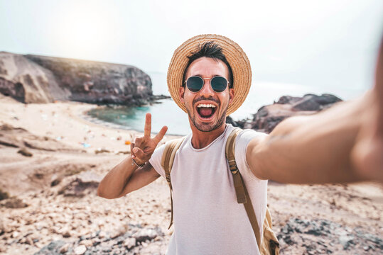 Young man with backpack taking selfie portrait on a mountain - Smiling happy guy enjoying summer holidays at the beach - Millennial showing victory hands symbol to the camera - Youth and journey