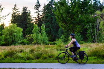 Naklejka premium Adventurous White Caucasian Adult Woman riding a road bicycle on a bike path at the famous Stanley Park in a modern city. Sunny Summer Sunset. Downtown Vancouver, British Columbia, Canada.