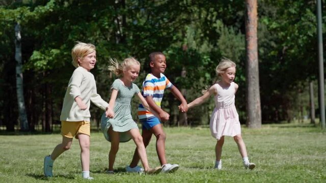 Tracking side view shot of group of happy multiethnic kids running on green grass holding hands. Diverse children hopping on lawn in park or woodland in slow motion and screaming with excitement