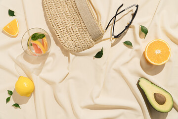 Straw clutch with sunglasses and coctail glass. Summer fresh citrus fruit and avocado with green leaves on a silky beige fabric.