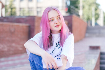 Positive pink-haired teenage hipster girl in a white T-shirt and blue jeans is using a smartphone on a summer day on the street of the city.Summer concept.Generation Z style.Social media concept.