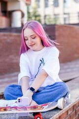 A pink-haired hipster teenage girl in a white T-shirt,jeans and with a skateboard is sitting on a city street on a summer day and using a smartphone.Generation Z style,social network,hobby.