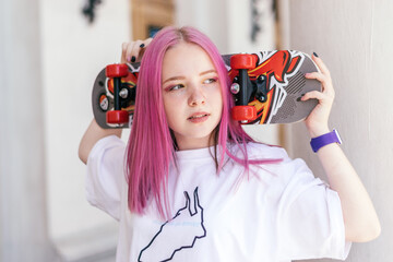 A pink-haired hipster teenage girl in a white T-shirt holding a skateboard on a city street on a summer day.Generation Z style,active lifestyle. © Tatyana