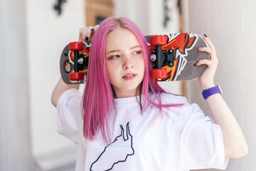 A pink-haired hipster teenage girl in a white T-shirt holding a skateboard on a city street on a...