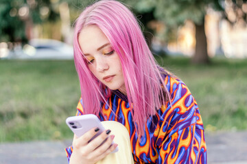 Pink-haired teenage hipster girl in a colorful bright T-shirt is using a smartphone on a summer...
