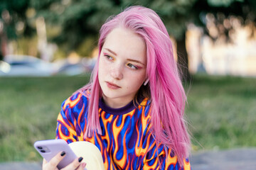 Pink-haired teenage hipster girl in a colorful bright T-shirt is using a smartphone on a summer...