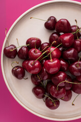 Overhead and closeup view of half a ceramic dish full of bright fresh appetizing cherries on a pink background.