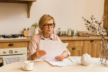 Middle aged mature senior woman holding paper bill or letter at home for making online payments on...