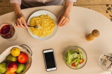 Mature senior woman in the kitchen preparing or eating tasty, homemade pasta in kitchen. Top view. Using mobile phone screen.