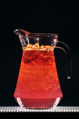 glass jug with red strawberry lemonade fizzy drink and ice on black background