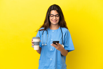 Young surgeon doctor woman isolated on yellow background holding coffee to take away and a mobile