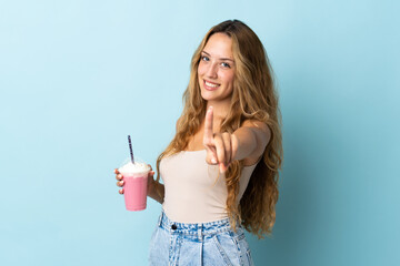 Young woman with strawberry milkshake isolated on blue background showing and lifting a finger