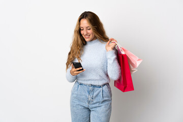 Young blonde woman isolated on white background holding shopping bags and writing a message with her cell phone to a friend