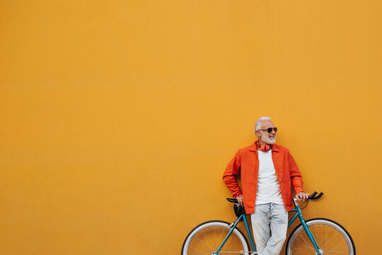 Smiling man in orange jacket and white shirt poses with bicycle. Attractive adult with white beard in light jeans posing on background of orange wall.. - Powered by Adobe